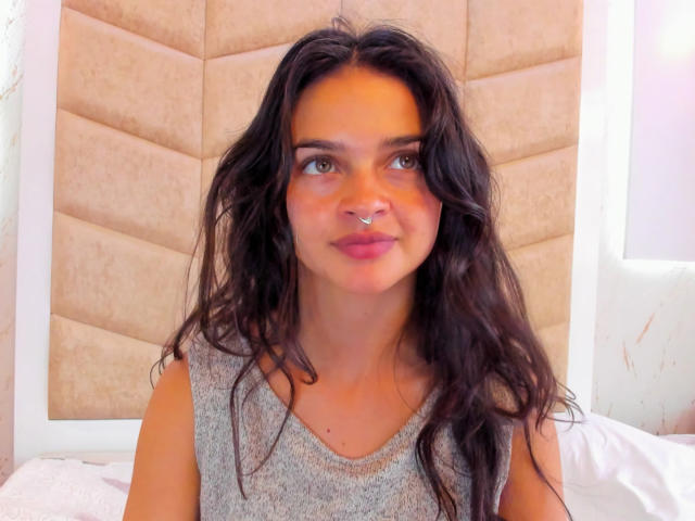LucyySky has wavy black hair, a silver nose ring, and is wearing a gray sleeveless top. Her green eyes are slightly upturned, and she's sitting on a white bed.