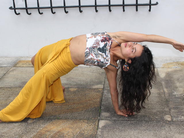 LucyySky is doing a yoga pose, back arching with arms stretched, wearing a floral crop top and yellow pants, her long curly black hair hanging down.