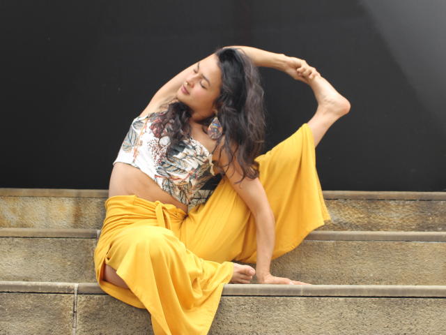 LucyySky is sitting on stone steps, wearing a yellow skirt and patterned top, with one leg raised and arm curved, showing off her flexible pussy and toned ass.