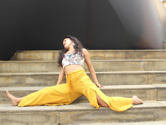 LucyySky is sitting on stone steps, legs spread wide, wearing a white floral crop top and yellow pants, with a tattoo on her right side, barefoot, eyes closed.