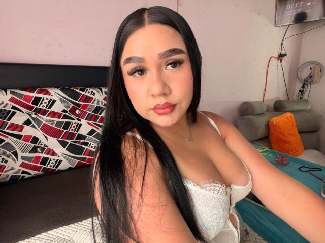 PaulettDelamour has long black hair, brown eyes, and red lipstick. She's wearing a white lace bra, showing her tits, and sitting at a table with a green surface.