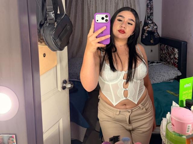 PaulettDelamour with long black hair, wearing a white lace-up bustier and beige pants, holding a purple phone, taking a mirror selfie, showing her tits and a slight belly.