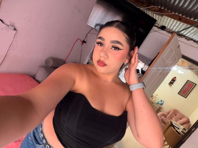PaulettDelamour with dark hair in a bun, black strapless top showing her tits, blue jeans, red lipstick, and a watch, taking a selfie with one hand on her head.