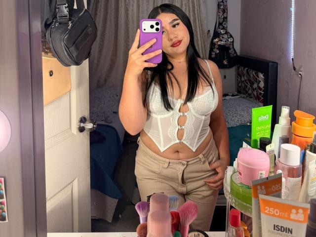 PaulettDelamour taking a mirror selfie, showing her white lace-up bralette that highlights her tits, paired with beige pants, standing in a bedroom with a cluttered vanity.