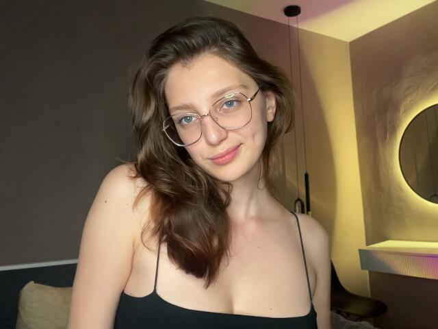 StaceyAverbach has light skin, blue eyes, wavy brown hair, glasses, and small tits in a black spaghetti strap top, smiling softly in a dimly lit room.
