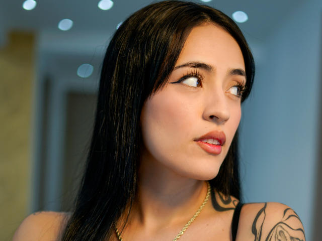 NicolleVanss with black hair, red lipstick, and tattoos on her right arm, wearing a black bra, hand on chest, necklace, and a confident smile, showing her tits.