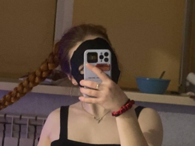 KatiRokisky with a black blindfold, holding a phone in front of her face, wearing a black tank top, red bracelet, and a braid. Boobs visible.