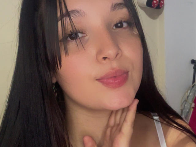 SarahGood with dark brown hair, slightly wet, looking at camera, pink lips, small nose, hand under chin, brown eyes, wearing white top, earrings visible, smiling gently.