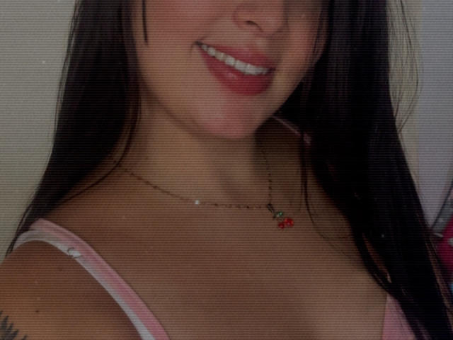 SarahGood's face with a smile, showing red lipstick and white teeth, wearing a pink and white striped top, necklace with red cherry pendant, and black hair.