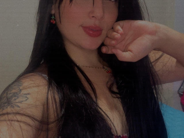 SarahGood with long black hair, pink lipstick, small tattoo on right shoulder, necklace, and earrings, showing her cleavage, holding her chin, wearing a low-cut top.
