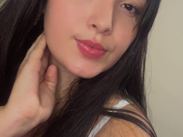 SarahGood with red lipstick, black hair, and a hand on her neck, showing off her cute face and slightly parted lips. Her tits are not visible.