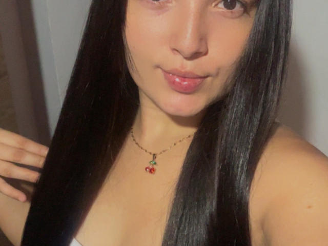 SarahGood with long black hair, pink lips, tan skin, wearing a red cherry necklace, slight smile, hands near neck, showing small tits, soft lighting, selfie style.