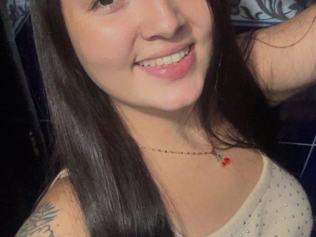 SarahGood has long dark hair, a cute smile with pink lips, and a necklace with a red flower. She's wearing a white top with small studs, showing a tattoo.