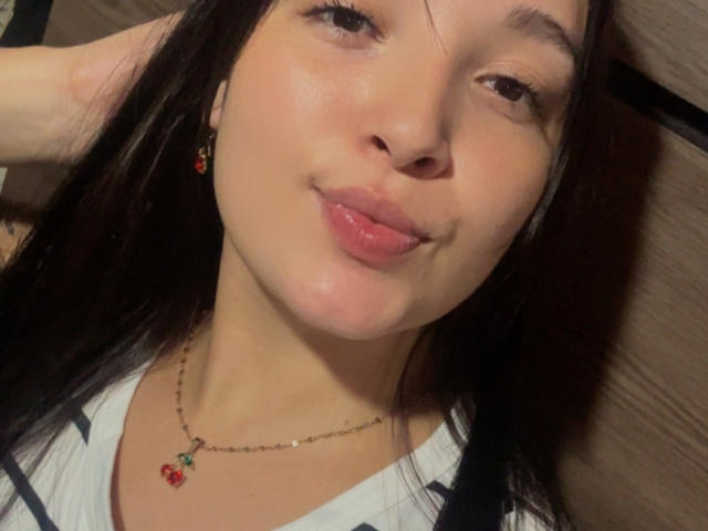 SarahGood with her tongue slightly out, wearing a red cherry necklace, black earrings, and a white shirt, has long dark hair and a cute smile.