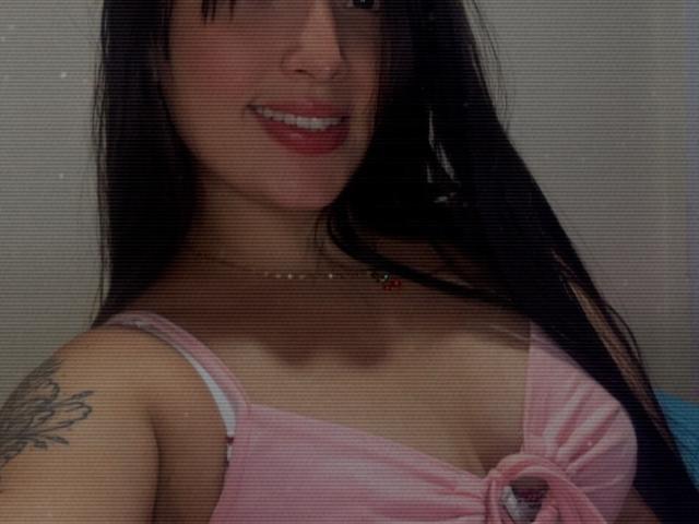 SarahGood with long black hair, pink top showing her tits, small tattoo on right arm, smiling with pink lipstick, necklace with red and green gem, grainy image.