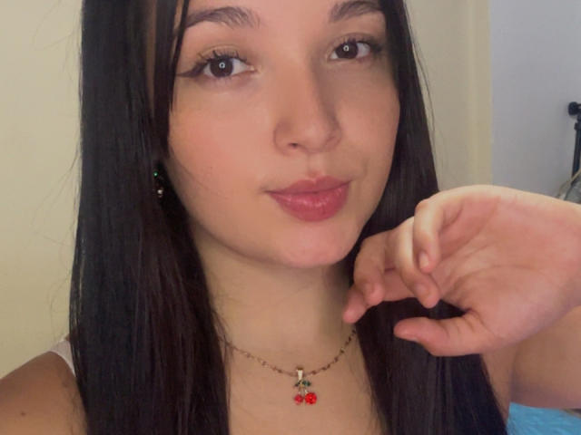 SarahGood has long black hair, light skin, and pink lips with a slight smile. She's wearing a cherry necklace and has her hand near her face.