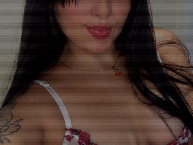 SarahGood with black hair, pink lips, wearing a red and white floral bra, showing her tits, a small red cherry necklace, and a tattoo on her right arm.