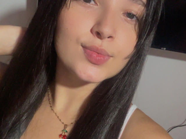 SarahGood has dark straight hair, pink lips, and light skin, wearing a white top and a red cherry necklace, with a slight smile.