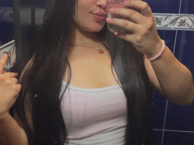 SarahGood with long black hair, white tank top, and necklace, taking a mirror selfie showing her tits, pouty lips, and pink wristband, holding phone with right hand.