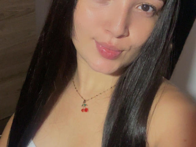 SarahGood has long black hair, pink lips, and a cute smile. She's wearing a necklace with a small red cherry pendant, and her bare shoulders are visible.
