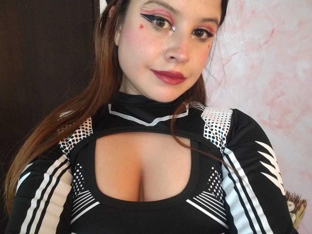 MelaniBecker with red lipstick, black and white striped top, cleavage, and pink eye makeup, smiling at the camera. Her brown hair is down, and she has a small red beauty mark.