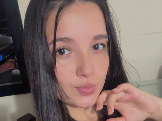 SarahGood with straight black hair, brown eyes, and glossy pink lips, slightly pouting, touching her chin with her right hand, in a casual selfie.