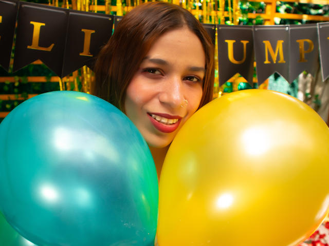 ValerieWoods with brown hair, smiling, holding yellow and blue balloons, nose pierced, pink lipstick, green and yellow background, festive atmosphere.