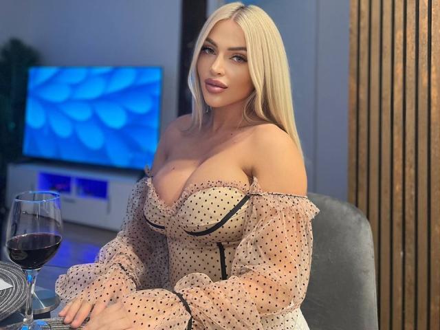 NadiaMarble with blonde hair, polka-dot dress, off-shoulder, revealing large tits, sitting at a table with wine, looking at camera. Her dress has black trim and ruffles.