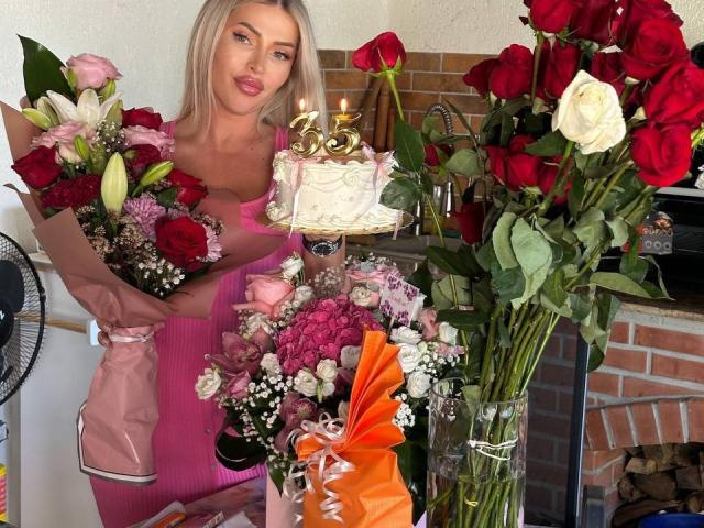 NadiaMarble with blonde hair, pink dress, holding bouquet of flowers, white cake with candles, and large bouquet of red roses, standing next to brick fireplace.