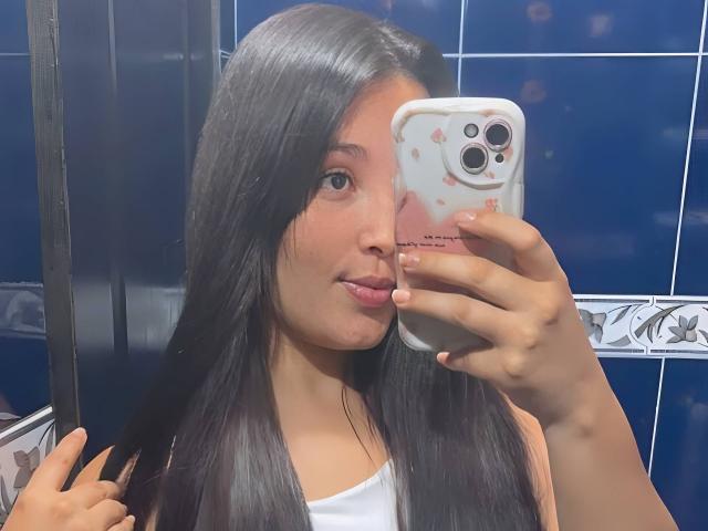 SarahGood with long black hair, holding a phone in front of her face, wearing a white tank top, in a blue-tiled bathroom, taking a selfie.