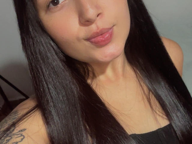 SarahGood has long black hair, light skin, and pink lips. She's smiling with a slight pout, wearing a black top, and has a tattoo on her right shoulder.