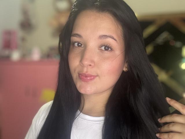 SarahGood has long black hair, brown eyes, and a slight smile with pink lipstick. She's wearing a white shirt, and her hand is touching her hair.