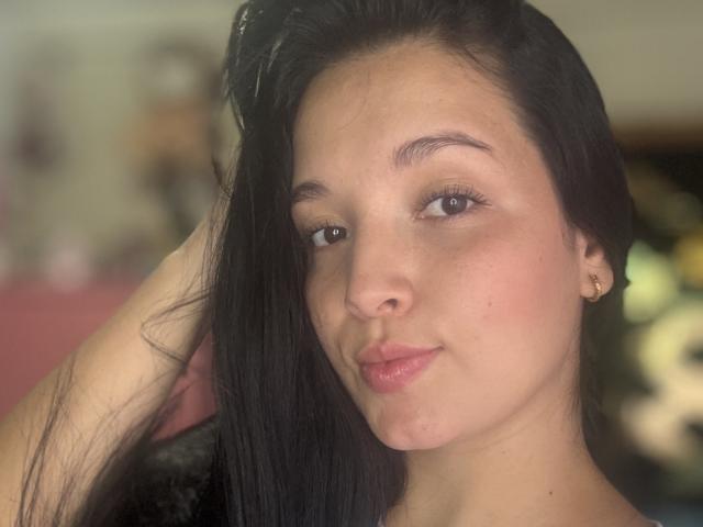 SarahGood has dark, straight hair, brown eyes, and pink lips, with a slight smile. She's wearing small gold earrings and touching her hair. Her skin is light.
