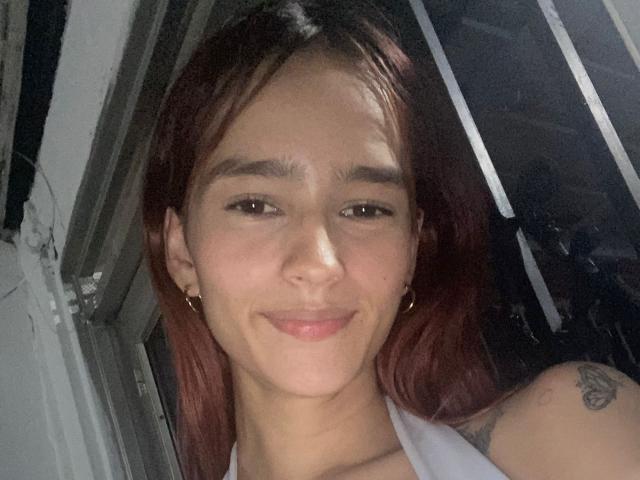 MelodyMansson has red hair, small tits, light skin, and a tattoo on her shoulder. She's smiling with brown eyes, wearing a white halter top, and gold earrings.
