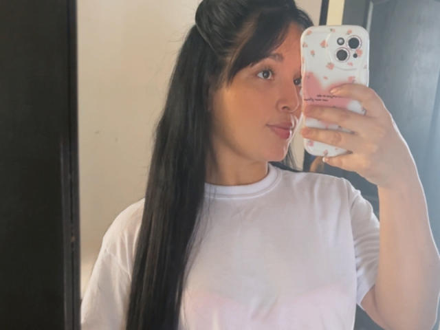 SarahGood with long black hair in a ponytail, wearing a white shirt, taking a mirror selfie with her phone, showing her tits slightly visible through the shirt.