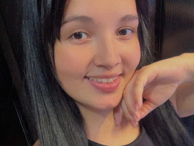 SarahGood has black hair, brown eyes, and a cute smile with pink lips. She's resting her chin on her hand, showing her soft, fair skin and relaxed expression.