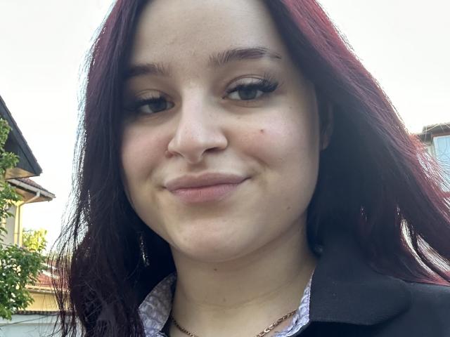 LucieRed has pale skin, dark red hair, and a small nose with a slight smile. She's wearing a black shirt over a patterned top, with a gold necklace.