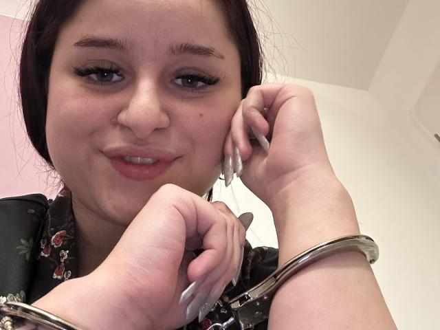 Lucie, avec ses cheveux bruns, porte un haut fleuri, a de longues ongles blanches et pointues et un bracelet argenté à son poignet. Elle sourit, tenant son téléphone à l'oreille..