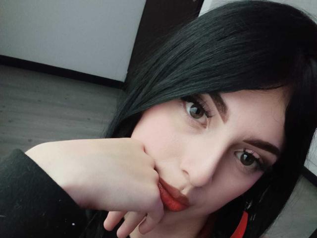 IsabellaaRossi with black hair, red lipstick, and pale skin, resting her chin on her hand, looking at the camera with green eyes, wearing a black top.