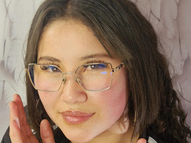 EmmaCruze has puffy pink cheeks, shiny pink lips, and is wearing gold-rimmed glasses with reflective lenses, her dark wavy hair and hoop earrings visible.