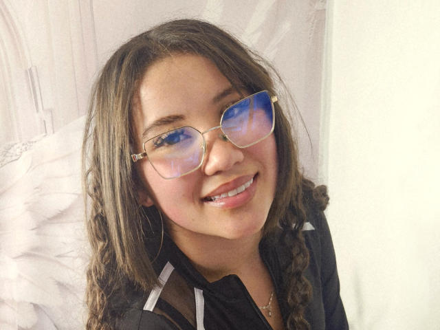 EmmaCruze with blue-tinted glasses, smiling, showing braces, wearing a black shirt with white trim, and brown braided hair, against a white background.