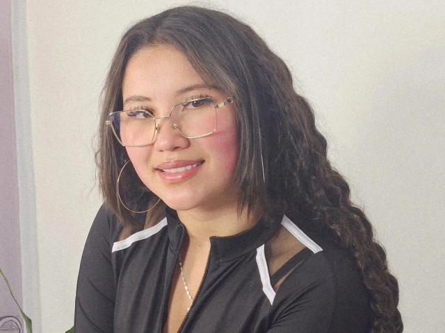 EmmaCruze has curly brown hair, glasses, pink blush, and a black top with white stripes, smiling with pink lips, wearing large hoop earrings, and a pearl necklace.