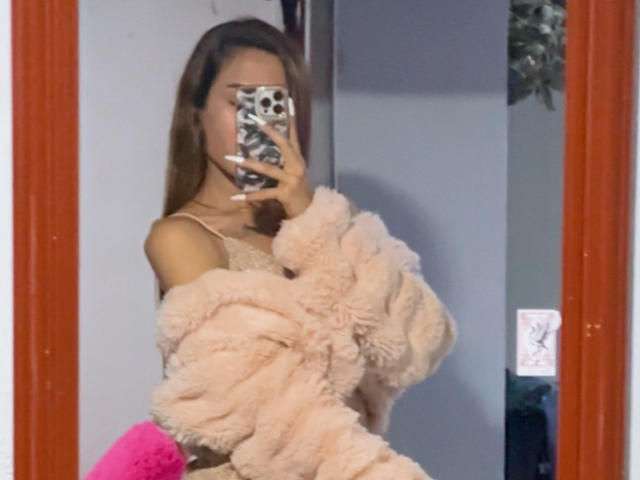 EmmaLym, con lunghi capelli castani, indossa una soffice maglietta rosa a spalle scoperte e sta scattando un selfie allo specchio con il cellulare in mano, mostrando leggermente il seno. Cornice dello specchio rossa..