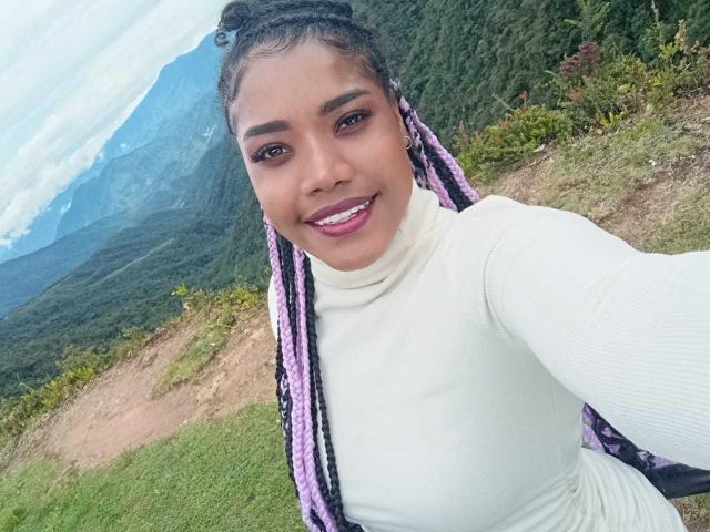 Caylin with purple and black braids, white turtleneck, smiling, showing her pearly white teeth, sitting on a grassy mountain, her cute brown eyes and full lips are visible.