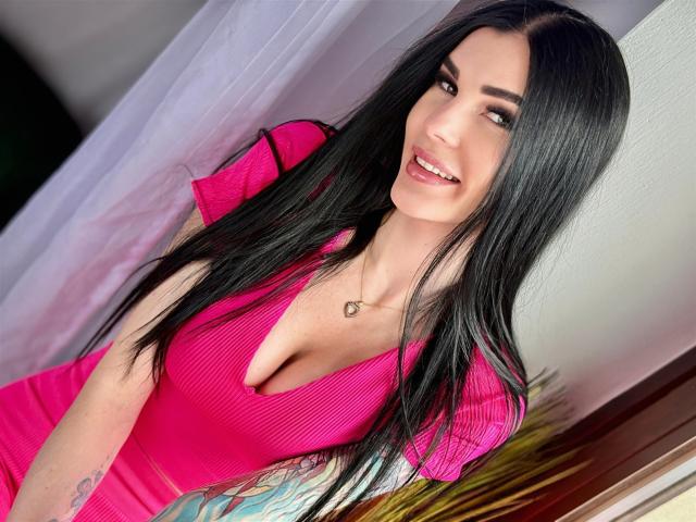 SofiaDream with long black hair, pink ribbed dress, heart necklace, smiling at camera, showing her big boobs, tattoo on right arm, standing in front of white curtain.