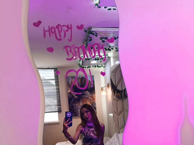 ValerieWoods taking a mirror selfie with purple pink glow, wearing a purple top, holding a phone, in a room with green garland and pink heart decorations. Boobs visible.