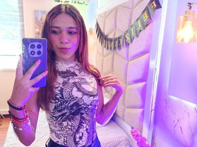ValerieWoods with long brown hair, wearing a white dragon print shirt, holding a blue phone, purple light, bed with white headboard, bangle bracelets, and a cute expression.