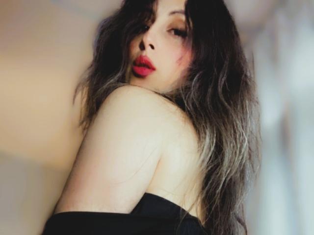 Sattine with messy, dark grey hair, red lipstick, and black off-shoulder top, looking over shoulder, showing bare shoulder and upper arm, slightly blushed face, sultry expression.