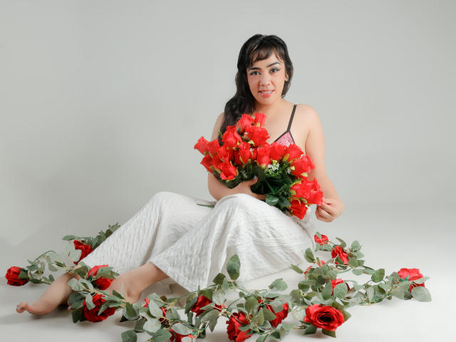 RayaHadley with red roses, holding a bouquet, sitting on white cloth, wearing pink bra, white skirt, surrounded by scattered roses, bare feet, smiling, black hair, pale skin.