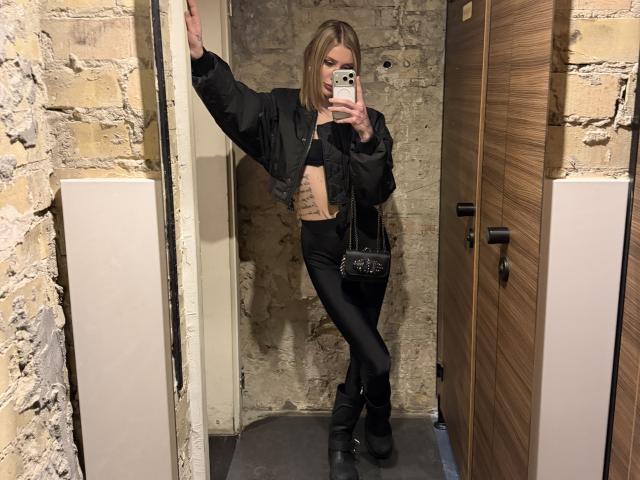 SensualMila with blonde bob, black jacket, black lace top, black leggings, black boots, holding phone, standing in brick-walled changing room, tattoo on right side of ribcage.